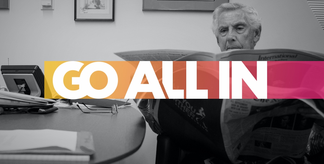 founder Al Golin reading the newspaper with gradient banner with words Go All In running across image.
