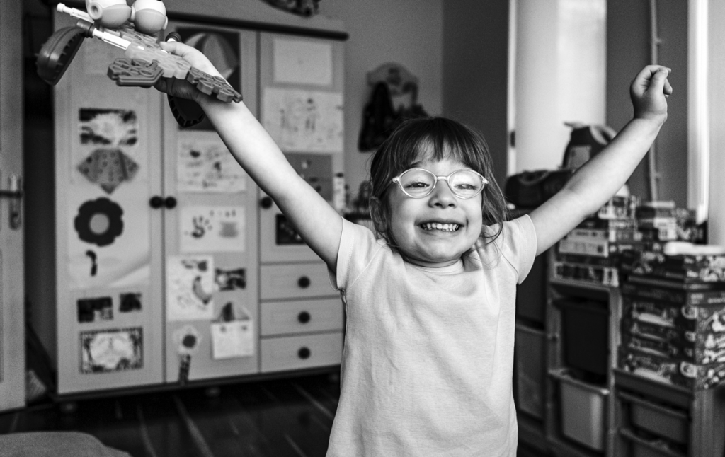 little girl with glasses smiling with arms spread wide open and toy in her hand. photo is black and white