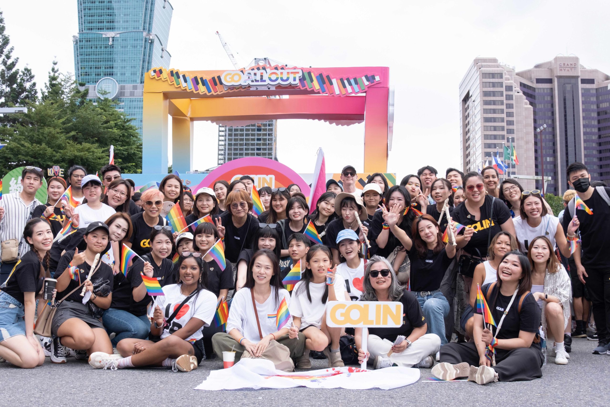 A large group of young people posing together at Taipei Pride. The group is gathered under a colorful rainbow-gradient arch with 'GO ALL OUT' text. Many participants are holding small rainbow flags and wearing black or white t-shirts. They're smiling and making peace signs, with some holding 'GO IN' signs.