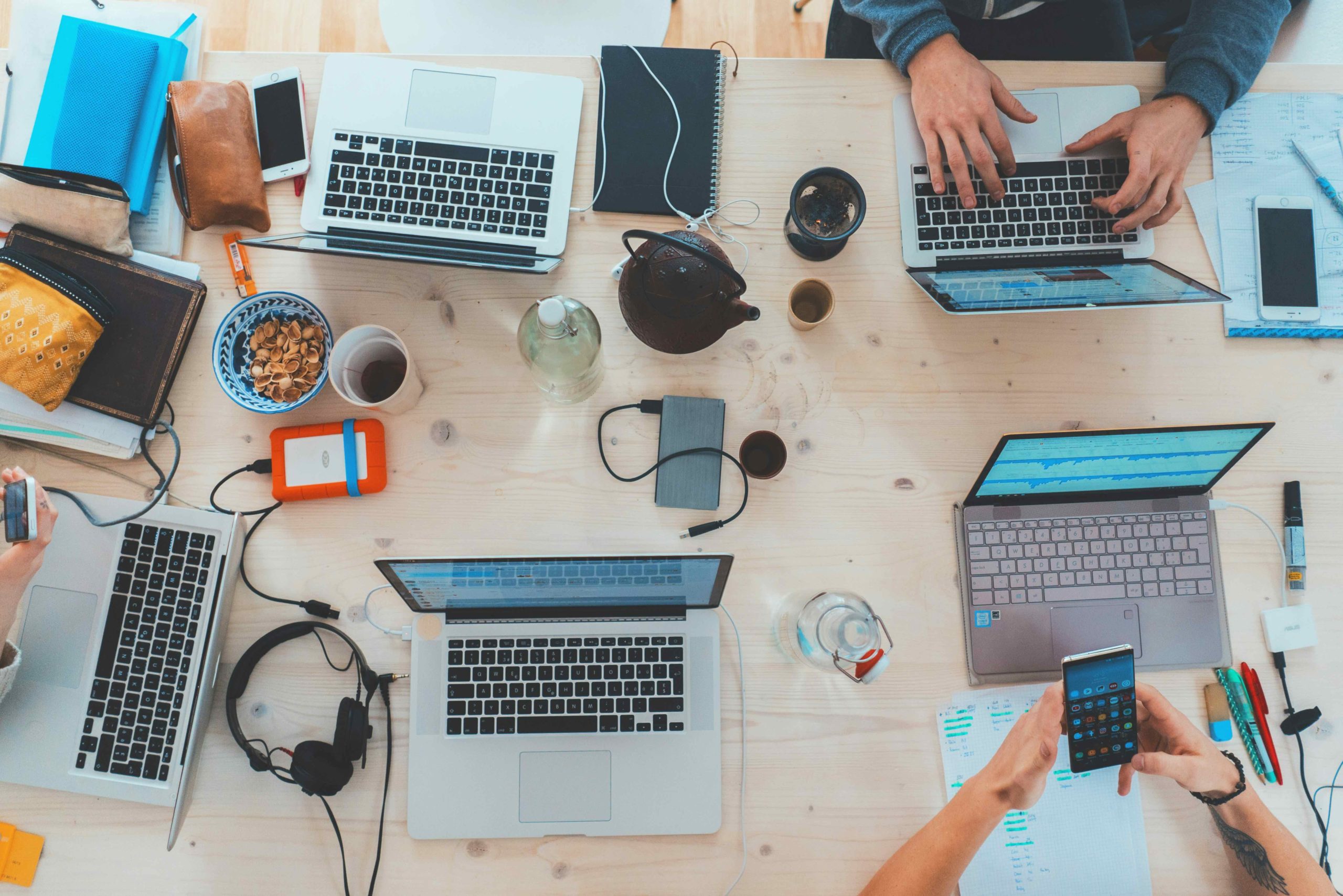 The image shows a cluttered desk filled with various electronic devices, laptops, and office supplies, suggesting a busy, technology-driven workspace. Different gadgets, cables, snacks, and stationery items are spread out on the wooden surface, indicating a multitasking environment.