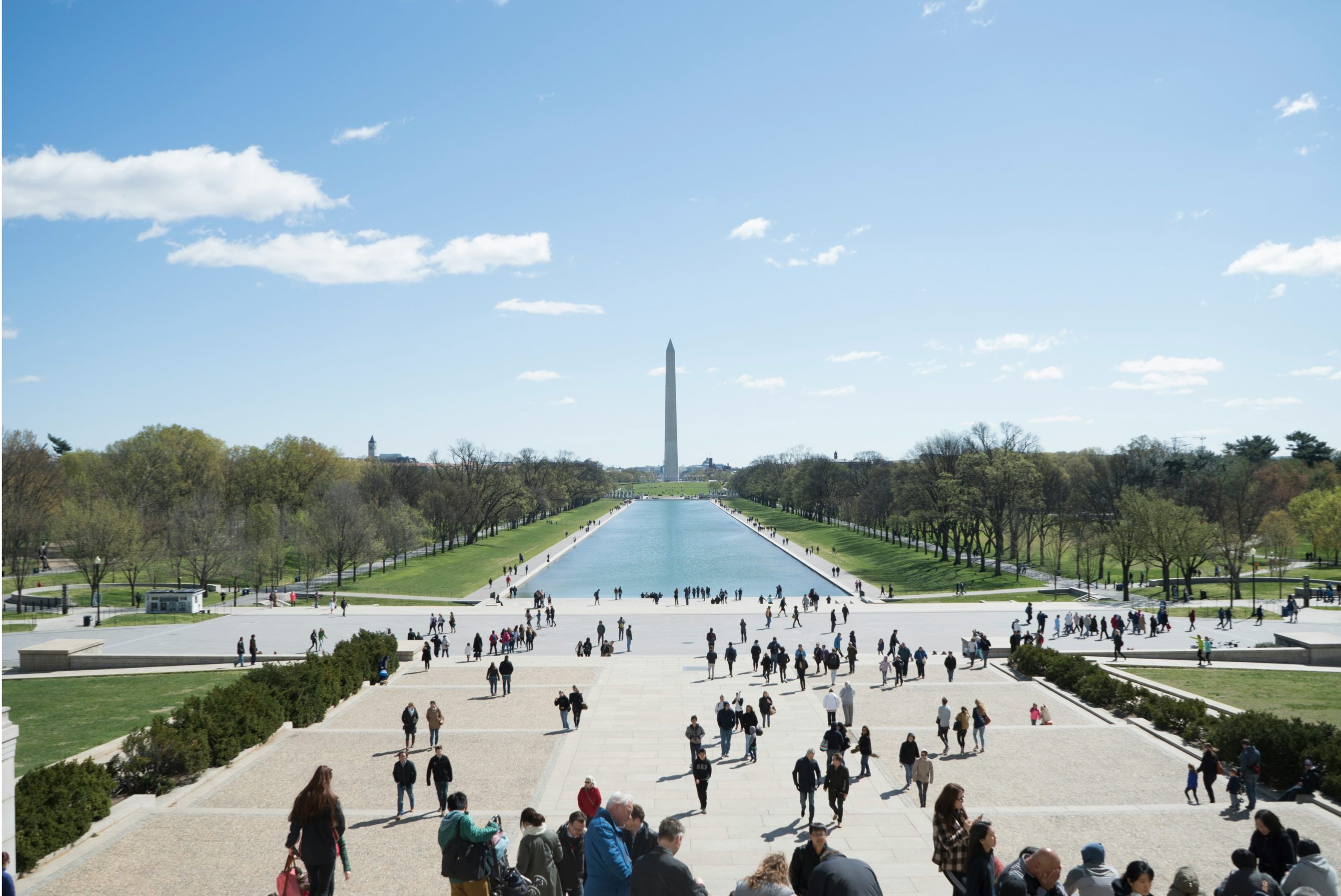 View from the steps of the Lincoln Memorial overlooking the National Mall in Washington, D.C., with crowds of people walking along the pathway and gathering near the Reflecting Pool.