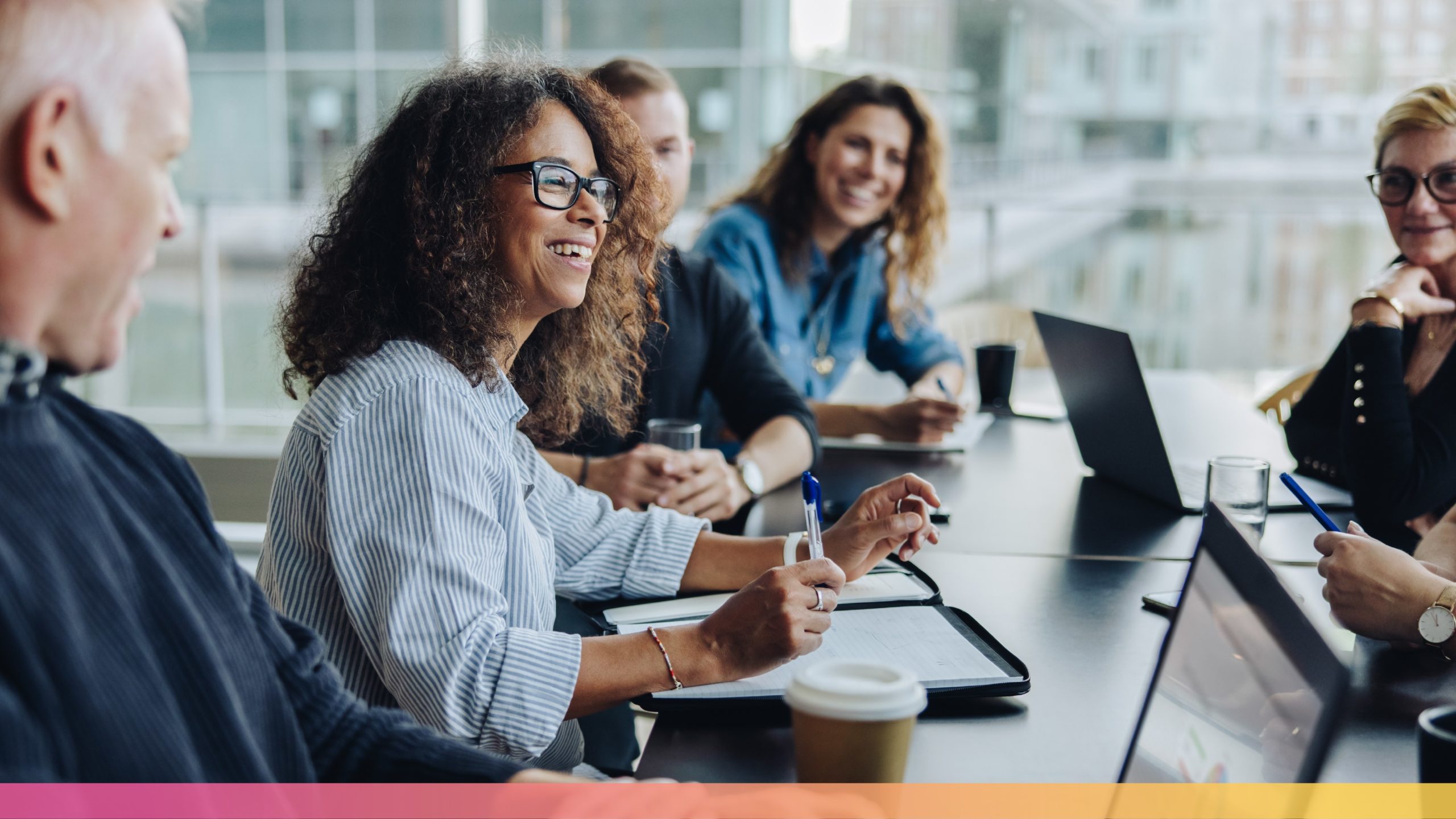 A diverse group of professionals share a genuine moment of laughter during a meeting in a light-filled, modern office space. Gathered around a sleek conference table with laptops and coffee cups, the team's natural camaraderie shines through, highlighted by the decorative gradient bar of pink, coral, and yellow tones at the bottom of the image.