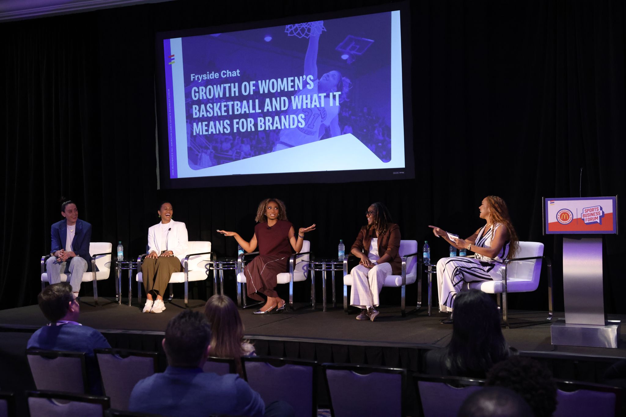 A panel discussion at a Sports Business Forum featuring five speakers seated on a stage discussing 'Growth of Women's Basketball and What It Means for Brands' as shown on the large screen behind them. The panel is titled 'Fryside Chat' with panelists in professional attire sitting in white chairs. A central speaker in a burgundy dress is gesturing while speaking, with other panelists listening attentively