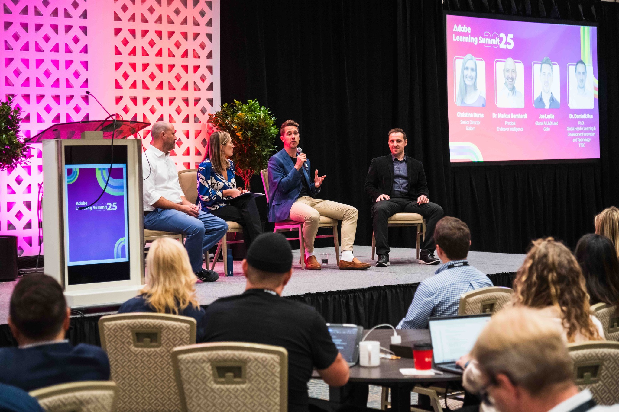 Four panelists sit on stage at Adobe Learning Summit 25, engaged in discussion before an audience. The stage features pink and coral geometric patterned backdrop panels, plants, and a large screen displaying speaker headshots. Attendees are seated at tables in the foreground watching the presentation.