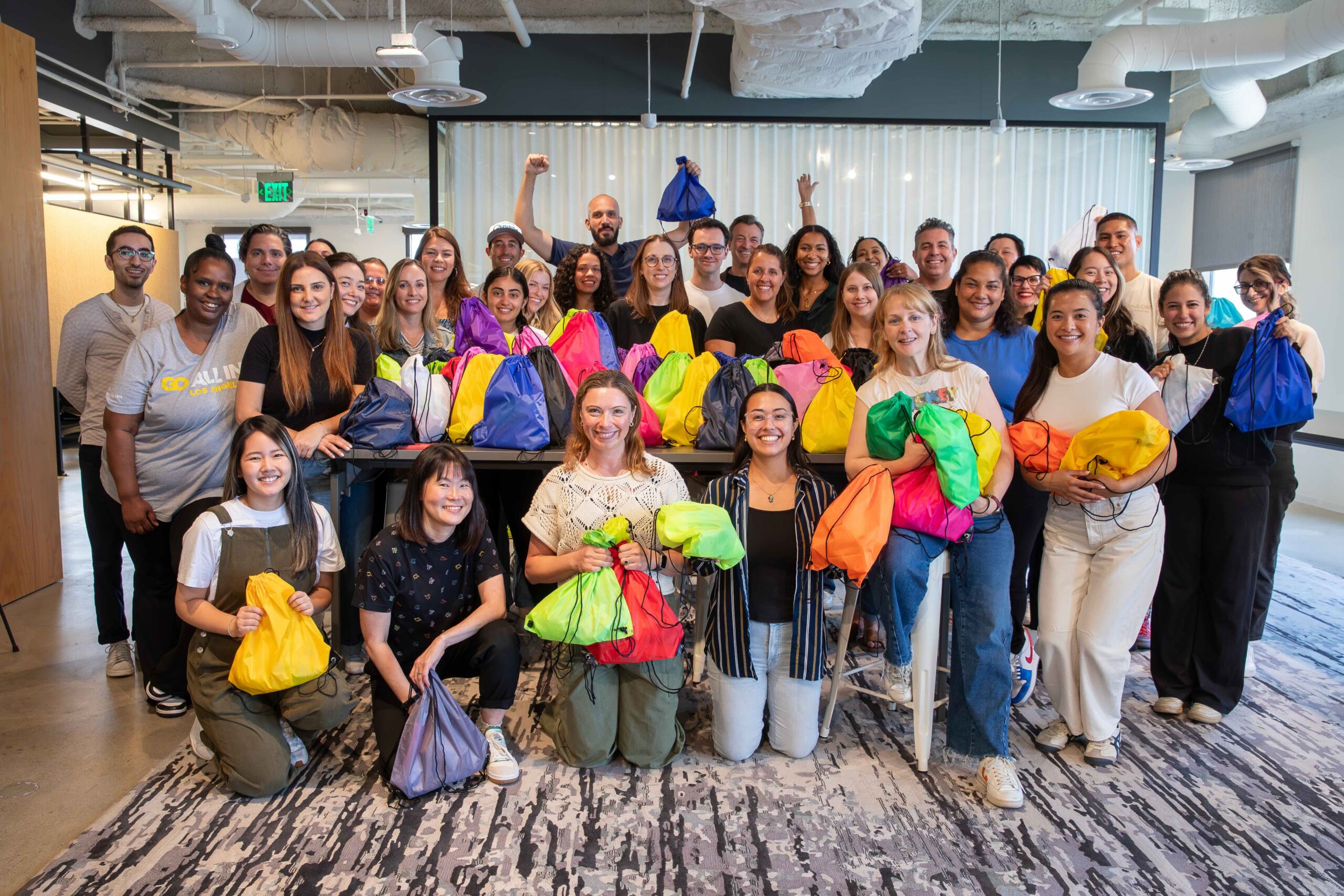 A large, diverse group of Golin employees gathered in a modern office space, smiling and holding colorful drawstring bags in bright shades of yellow, green, pink, orange, blue, and purple. The team is arranged in rows with some standing and others kneeling in front, celebrating together in a casual, energetic atmosphere with exposed ceiling elements and office furniture visible in the background.